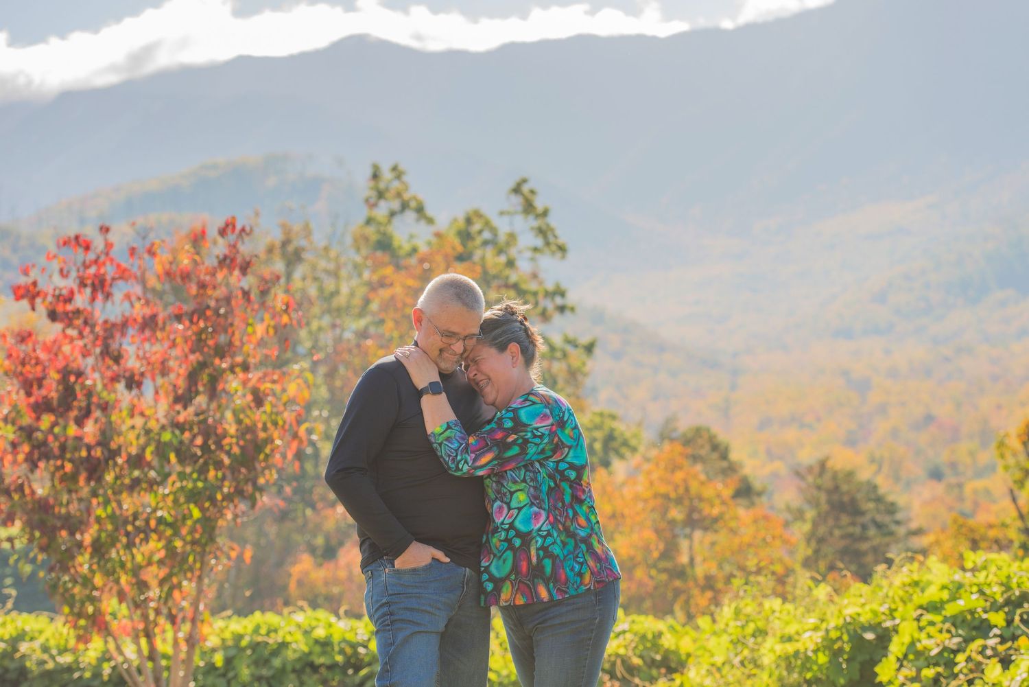 A couple embraces on a scenic overlook during autumn with colorful fall foliage and mountains in the background by Gatlinburg Photographers.
