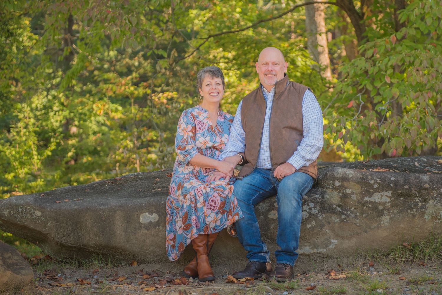 An anniversary  couple sits together on a large rock outdoors during golden hour with lush greenery in the background in Sevierville.