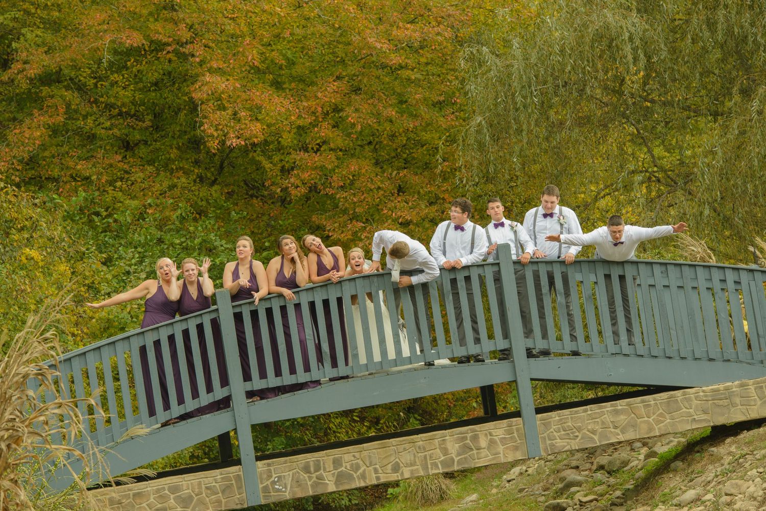 A wedding group lies in a row on a curved footbridge with autumn foliage in the background in Pigeon Forge TN.