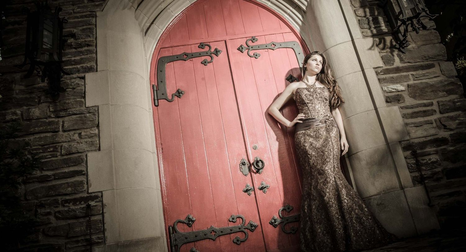 A figure in a sparkly gown poses beside a massive red arched door with ornate black metal hinges at a stone castle.