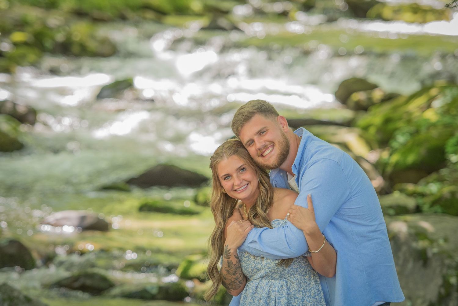 A happy couple embraces by a rushing mountain stream after proposal surrounded on a sunny day by Sevierville TN Photographers.