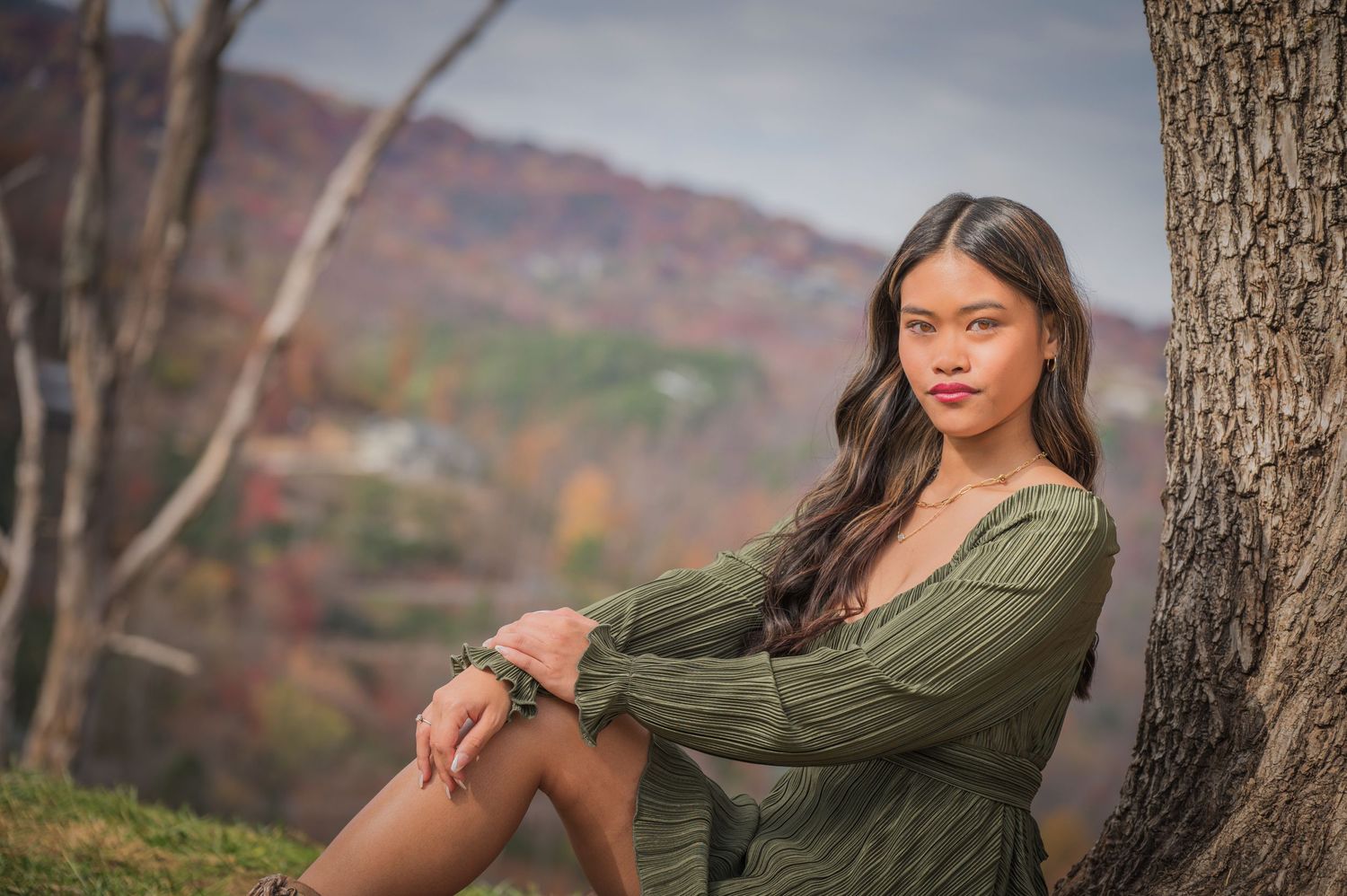 A figure in a green dress poses thoughtfully against a tree trunk with autumn foliage and mountains in the background.