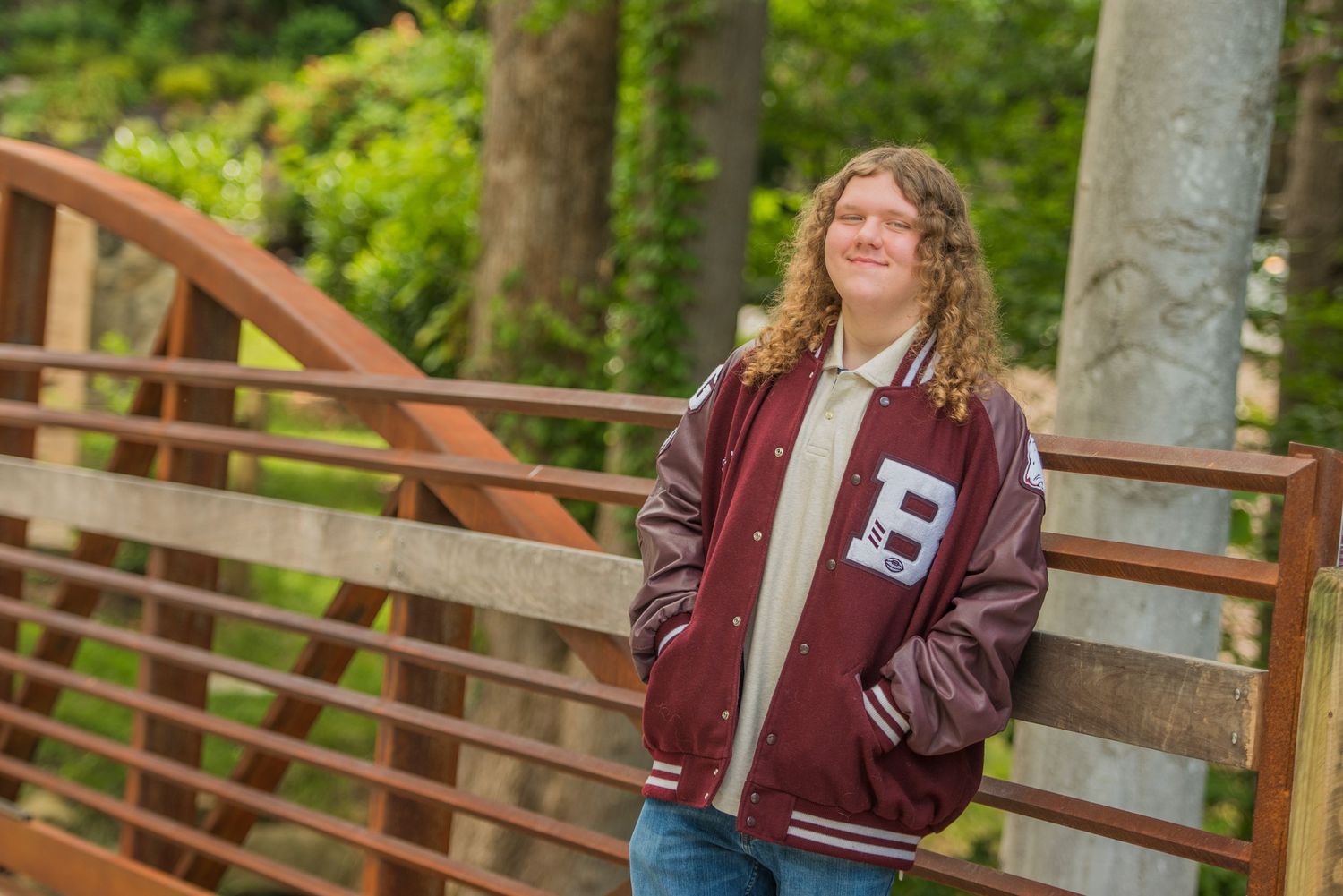 Student wearing a maroon varsity letter jacket poses on a wooden bridge surrounded by lush greenery.
