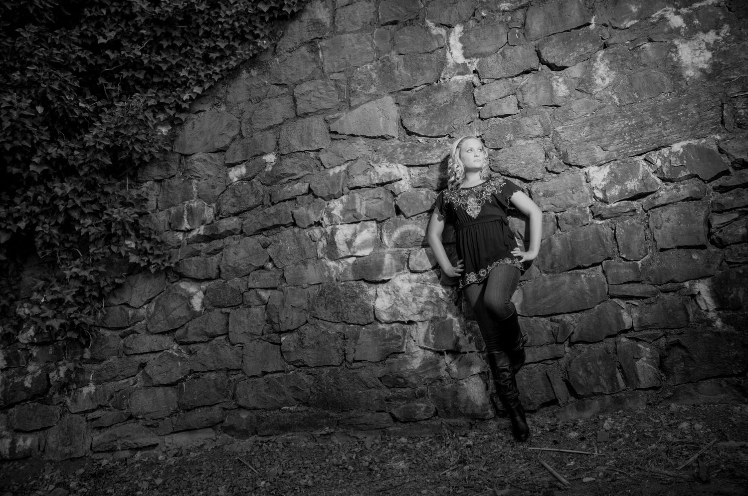 Dramatic black and white photo of a person posing against a rustic stone wall with a moody artistic aesthetic.
