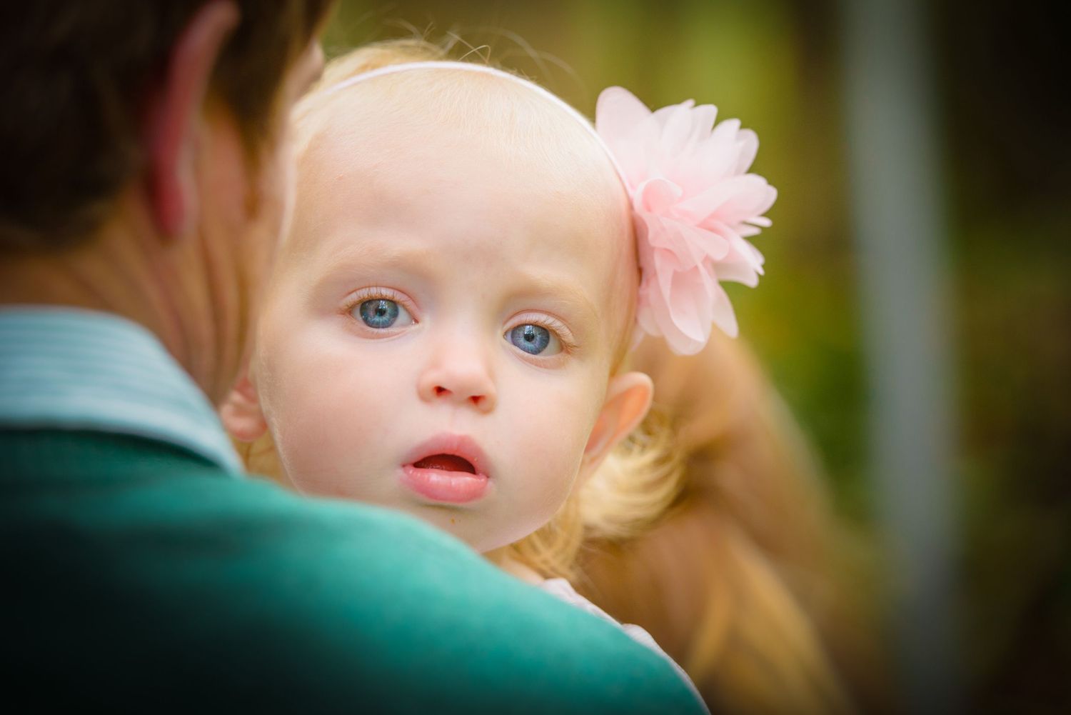 A small child with light blonde hair and bright blue eyes peeks over someone's shoulder wearing a pink flower accessory in Gatlinburg TN.