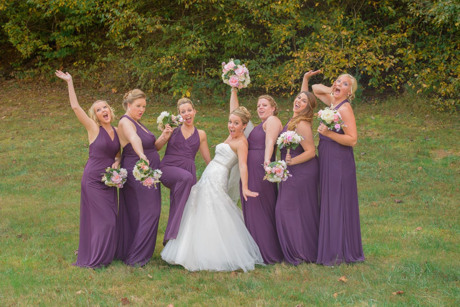 A group in purple bridesmaid dresses and a bride in white celebrate joyfully on a grassy lawn with bouquets held high in Pigeon Forge TN.