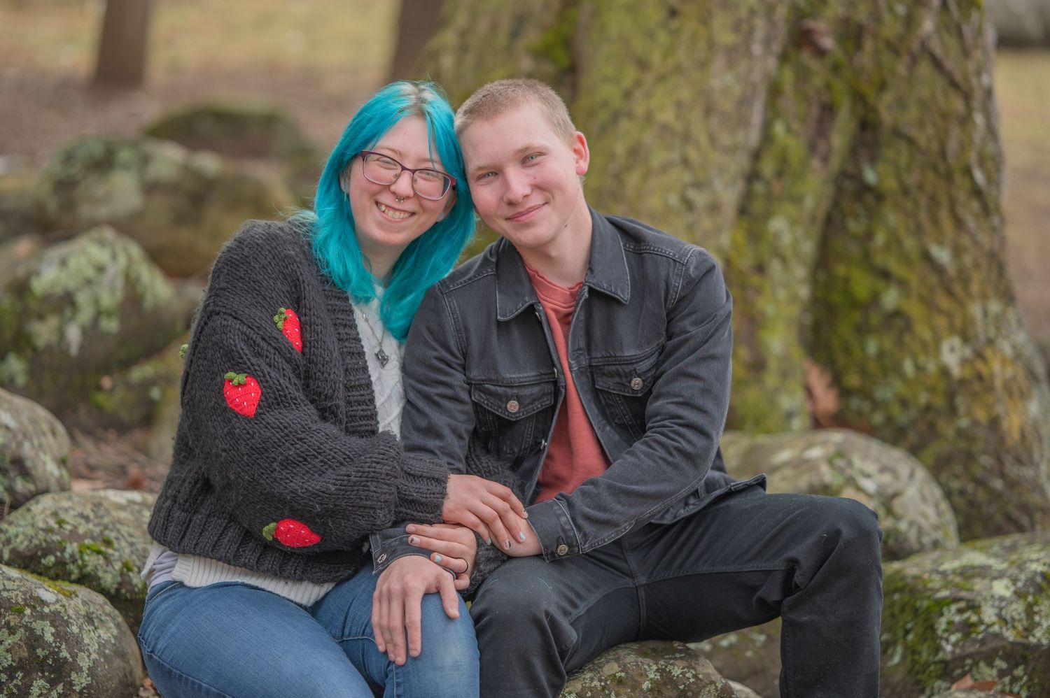 A couple sits together on mossy rocks by a large tree trunk, with one person wearing teal hair and a red heart sweater.