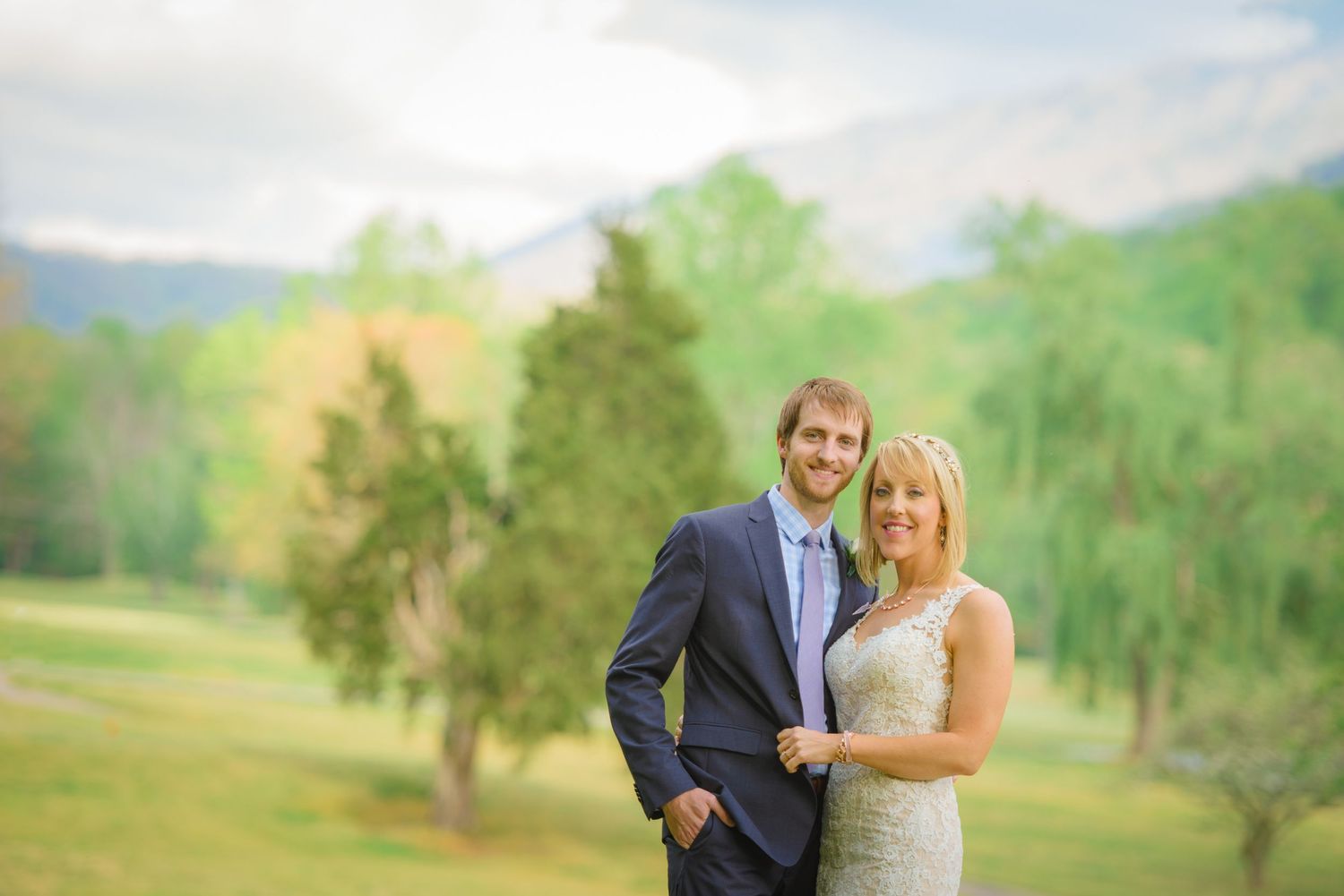 A couple in wedding attire embrace while standing in a scenic mountain meadow with green trees in the background by Gatlinburg Weddings.
