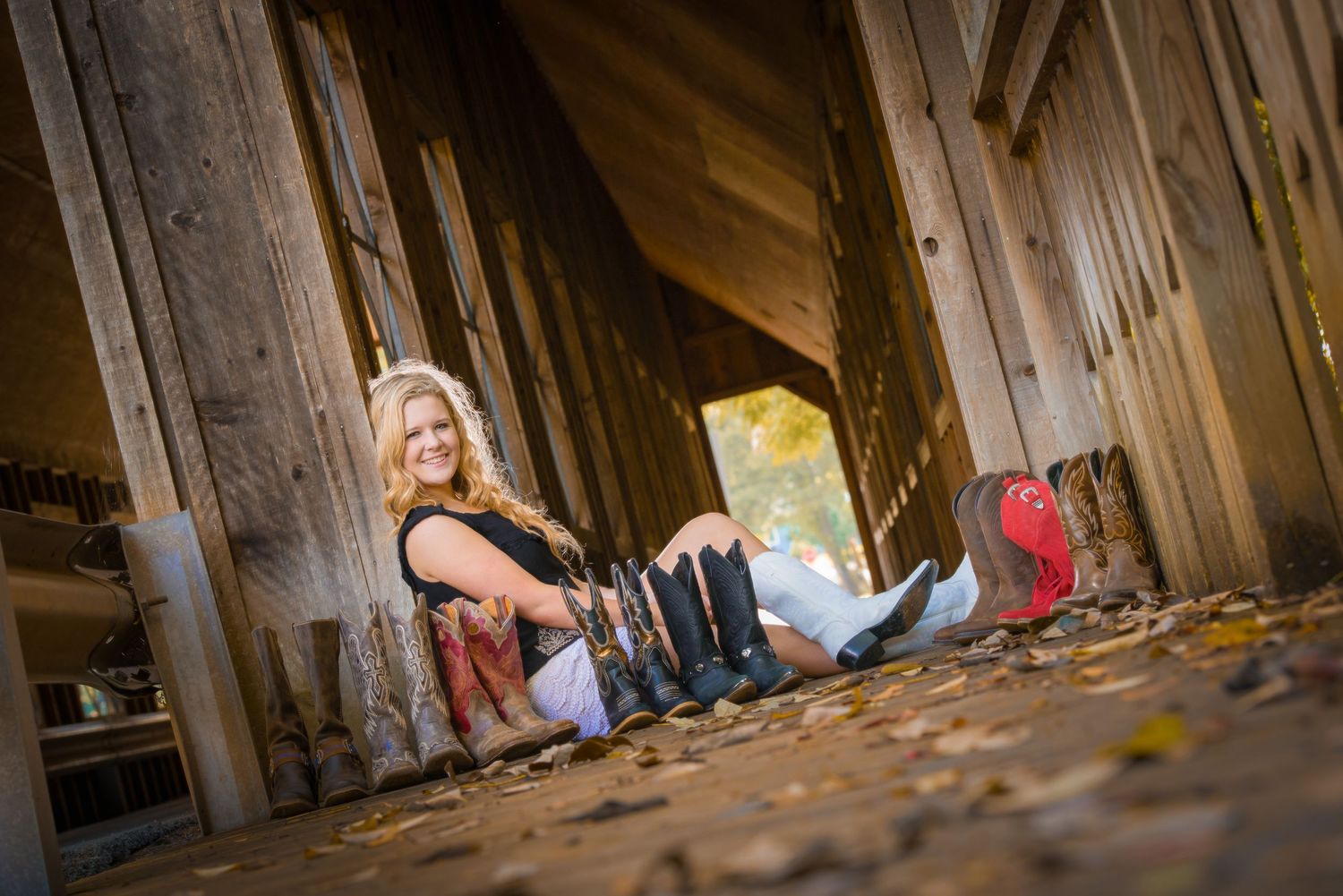 Someone sits casually on fallen autumn leaves under a rustic wooden bridge or covered structure in warm lighting.