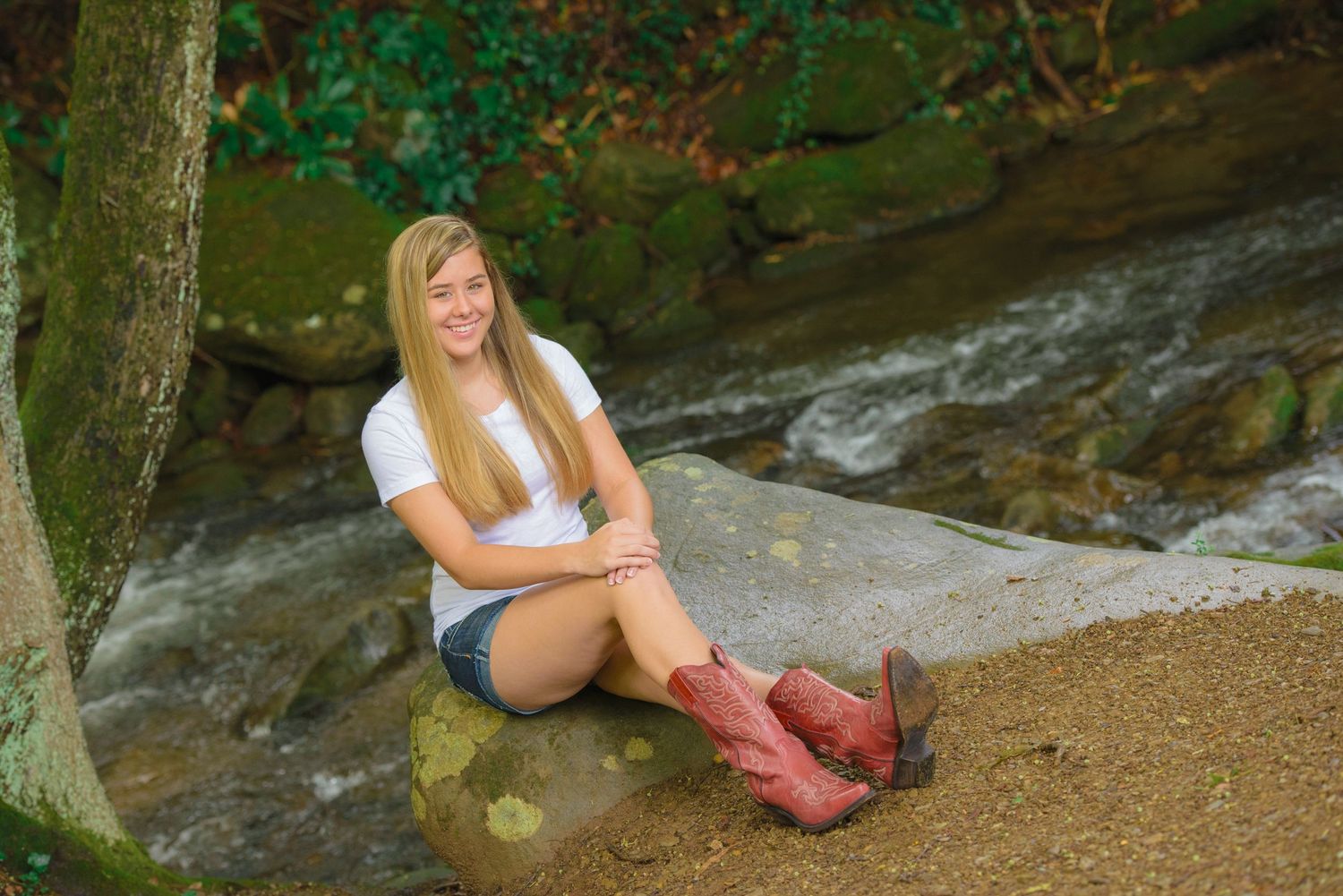 Someone in a white t-shirt and brown boots sits on a rock by a flowing stream in a forest setting.