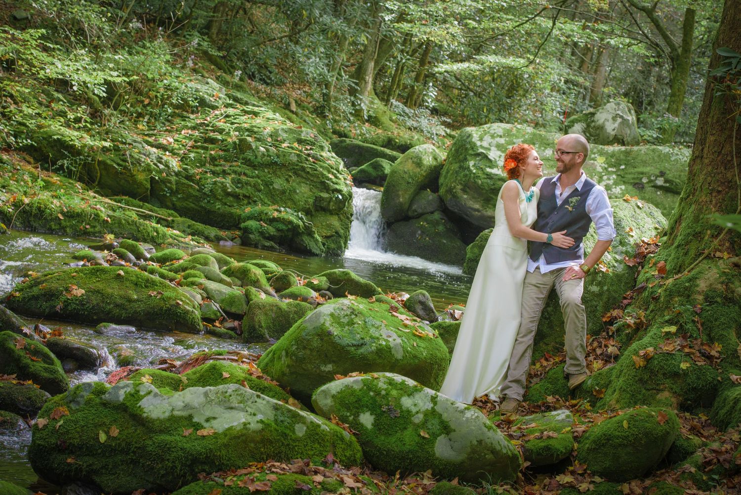 Two people in wedding attire embrace near a cascading waterfall surrounded by mossy rocks and lush forest greenery by Gatlinburg Weddings.