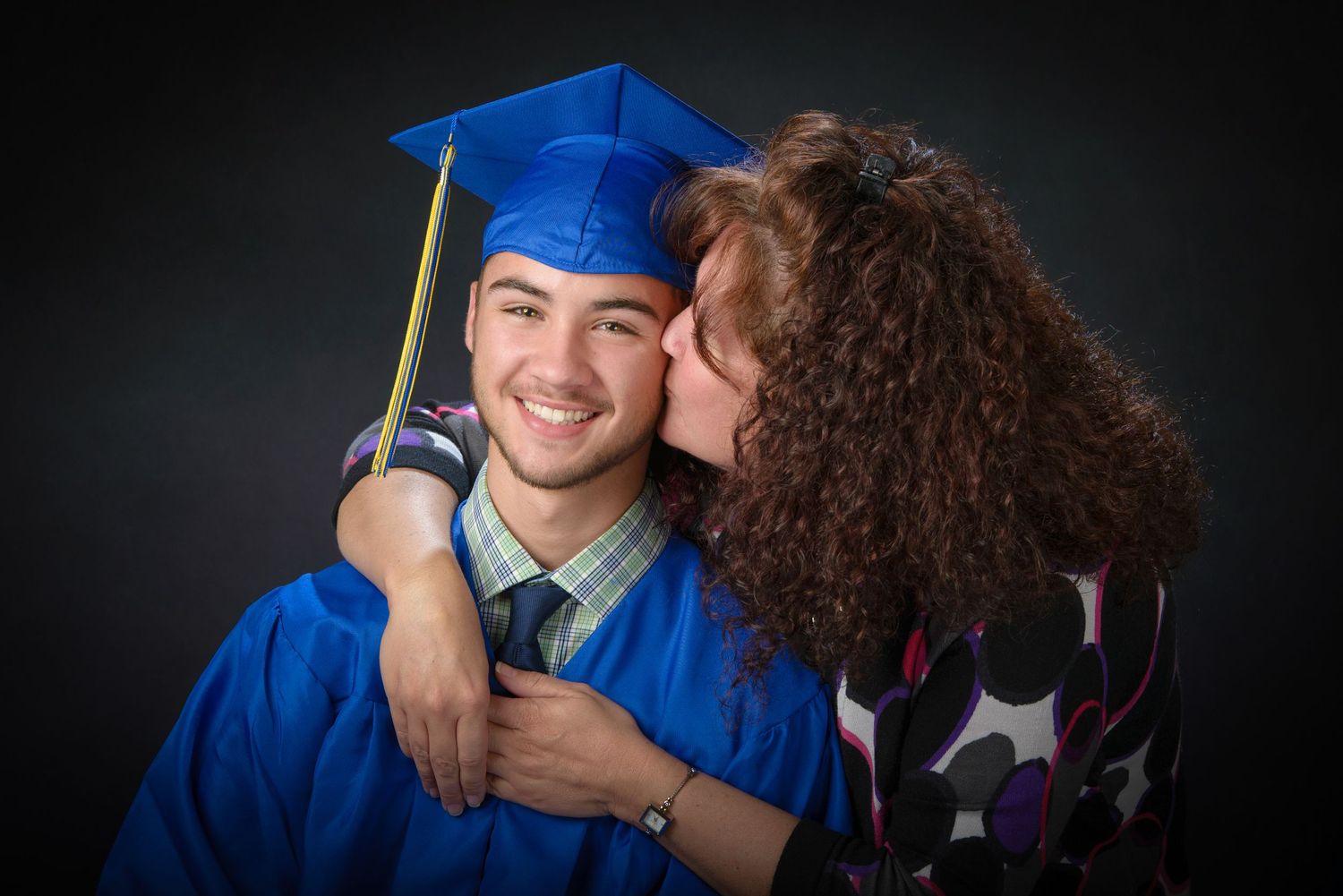 A graduate in a bright blue cap and gown receives an affectionate moment during a graduation photo session against dark backdrop.