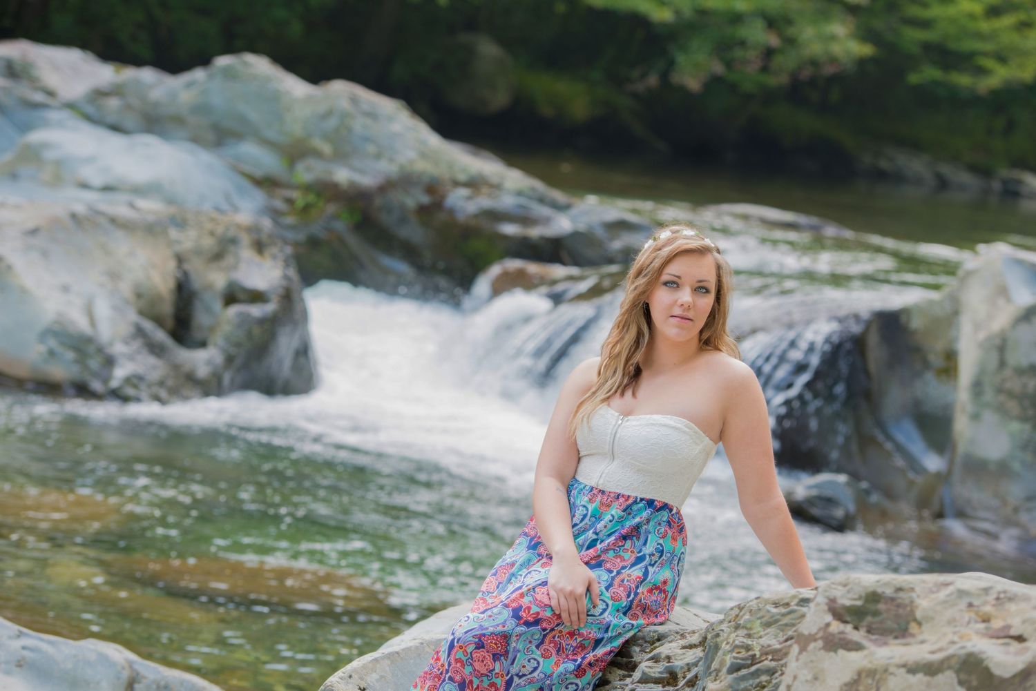 A person in a floral sundress sits gracefully on rocks beside a cascading waterfall in a serene forest setting.