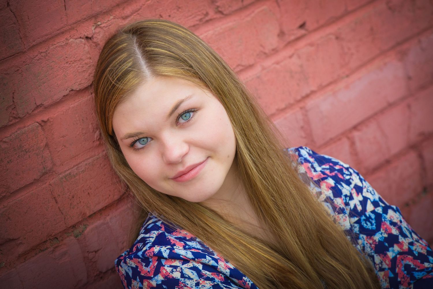 A portrait of someone with long blonde hair wearing a blue floral print dress against a red brick wall.