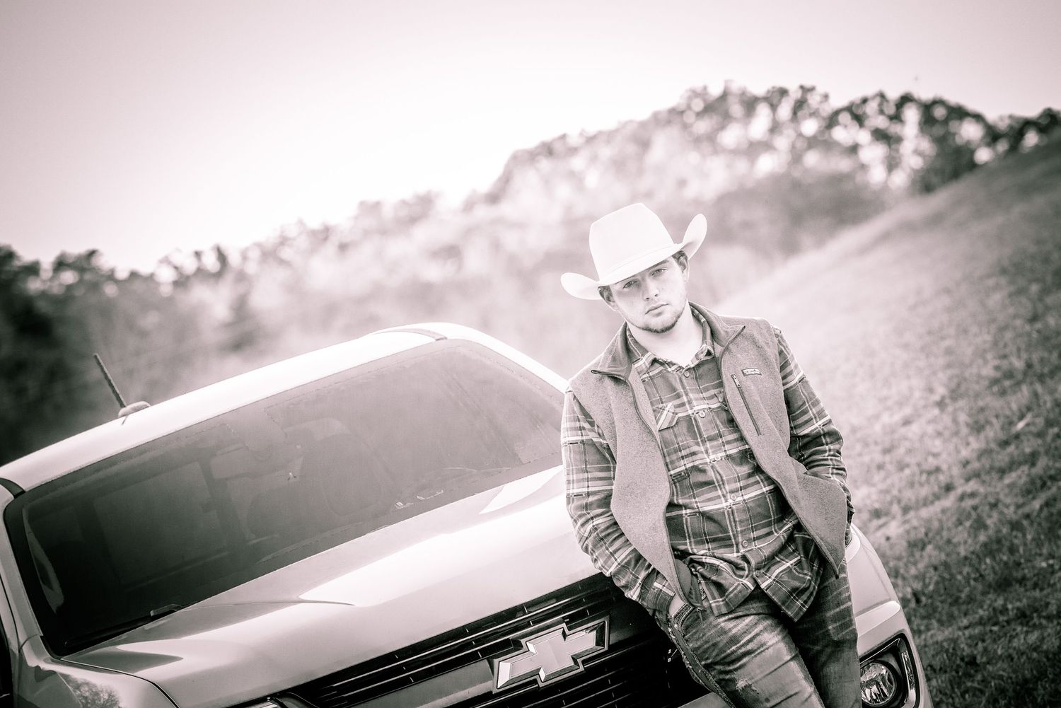 A black and white portrait of someone in a cowboy hat and denim jacket leaning against a car with mountains in the background.