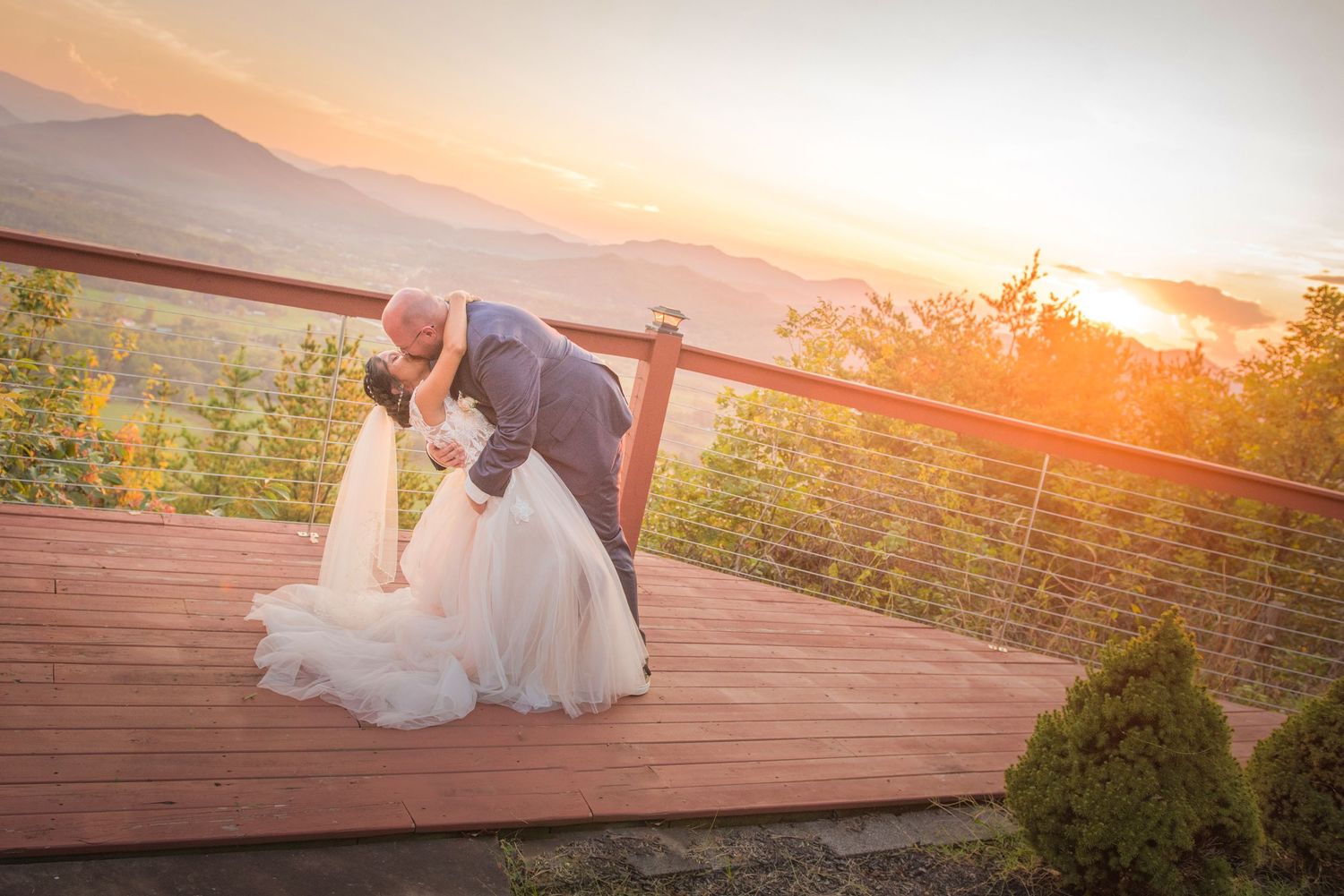 A couple sharing a romantic dip on a wooden deck overlooking mountain views during a golden sunset.