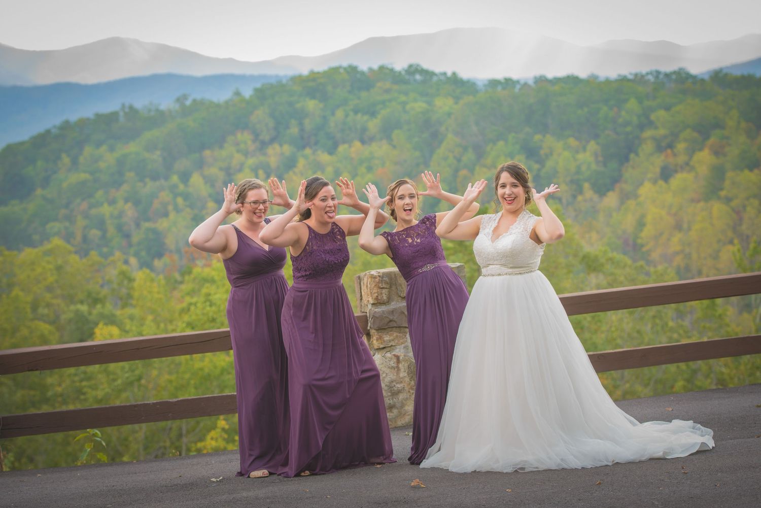 A bride in white and bridesmaids in purple dresses pose playfully on a mountain overlook with forest views in background in Pigeon Forge.