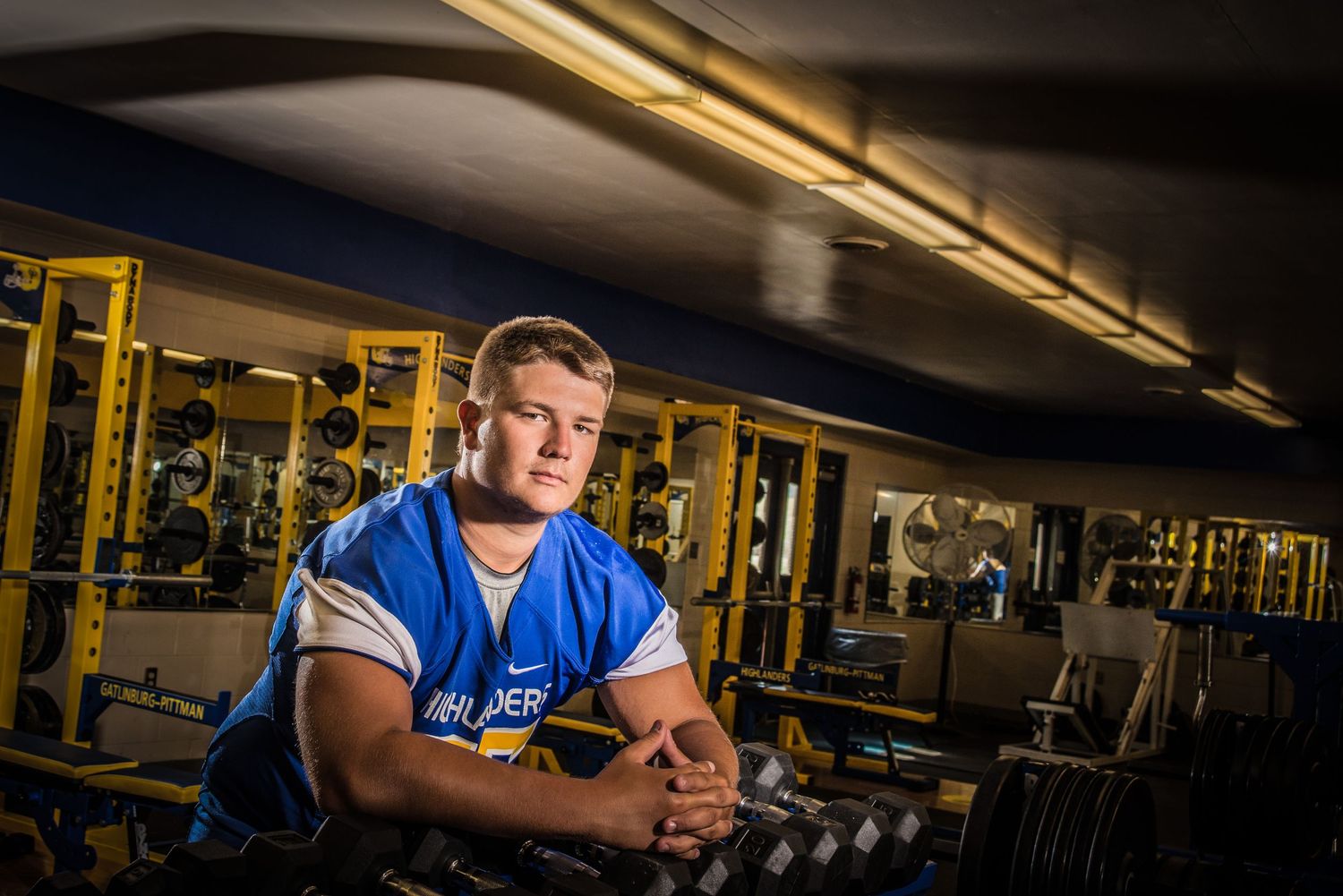 Athlete in blue shirt sits in empty gym with dramatic lighting and exercise equipment in background.