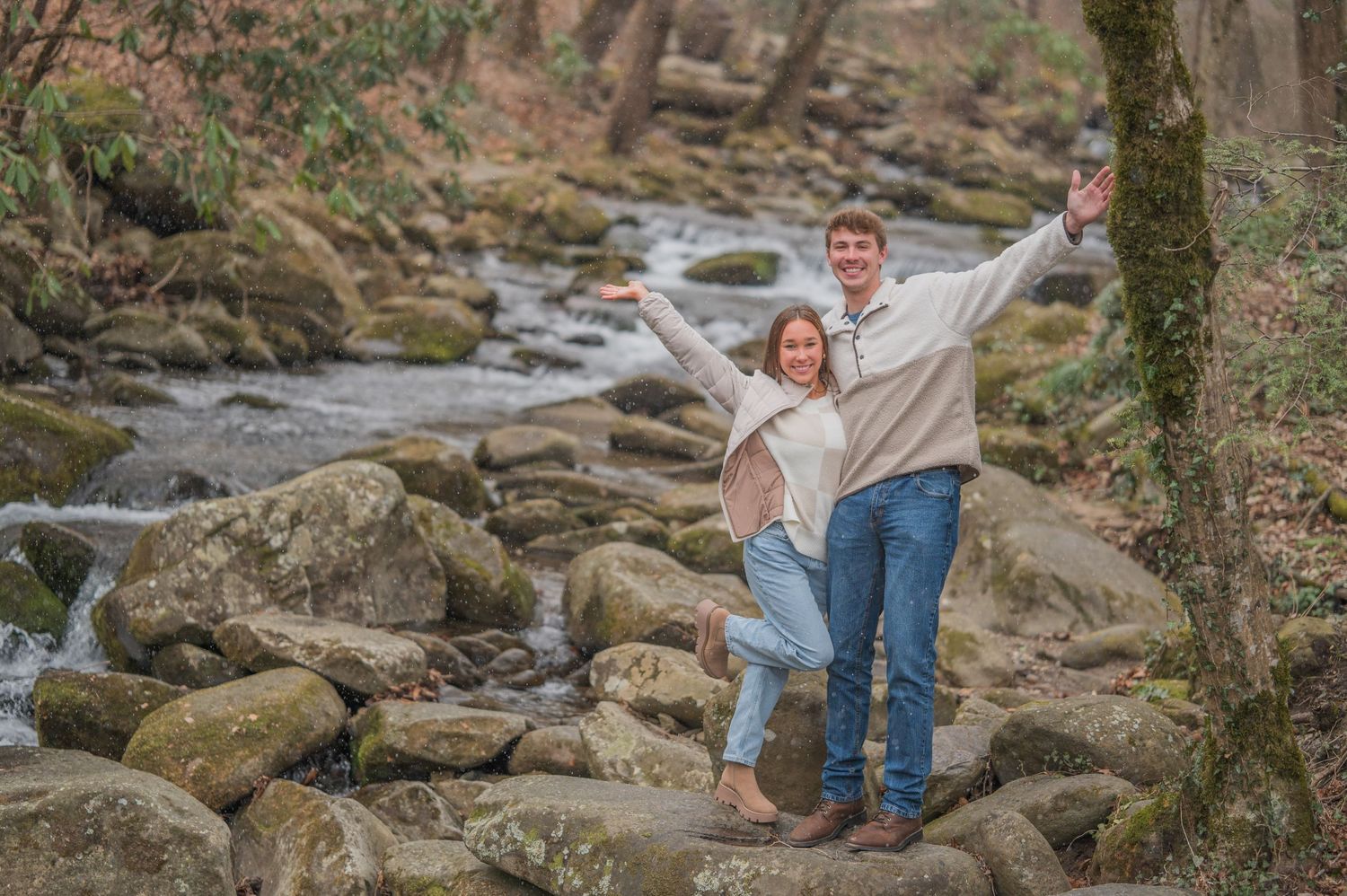 A joyful pair stand on rocky terrain by a flowing stream in the forest with arms raised in celebration by Pigeon Forge Photographer tn.