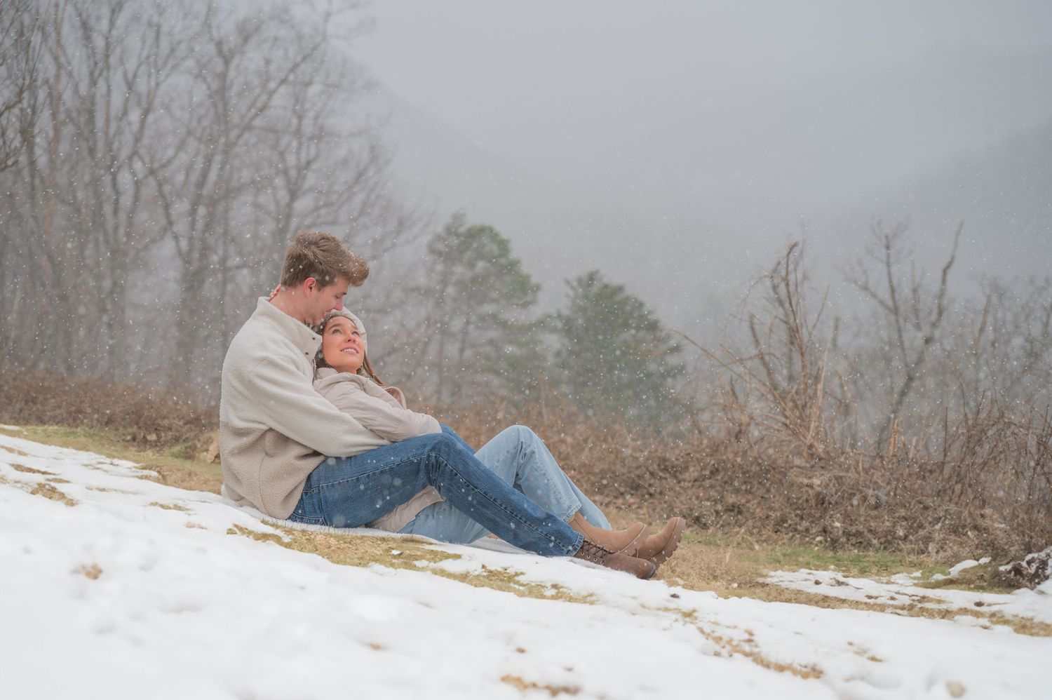 An engagement couple sits thoughtfully on a snowy hillside wearing jeans and a light jacket on a foggy winter day in Pigeon Forge.