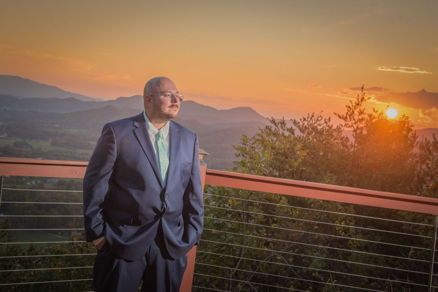 Groom in a suit stands on an outdoor deck against a stunning mountain sunset background by gatlinburg wedding photographers tn.