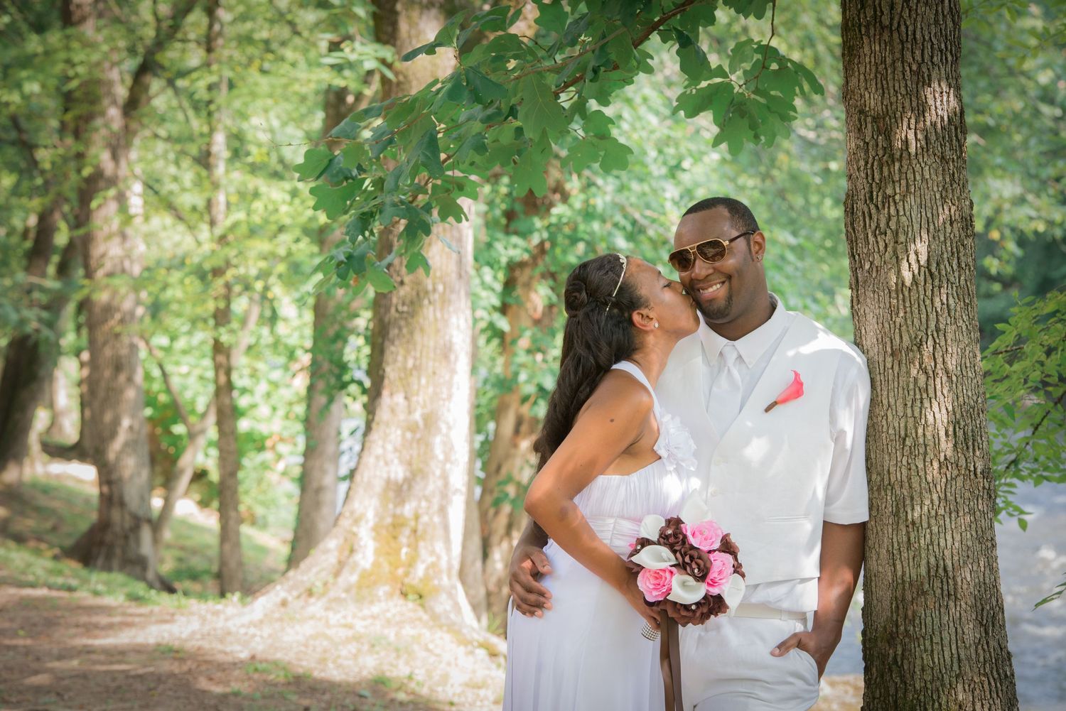 A couple in white clothing embrace while holding a pink bouquet in a sunlit wooded setting in Pigeon Forge TN.