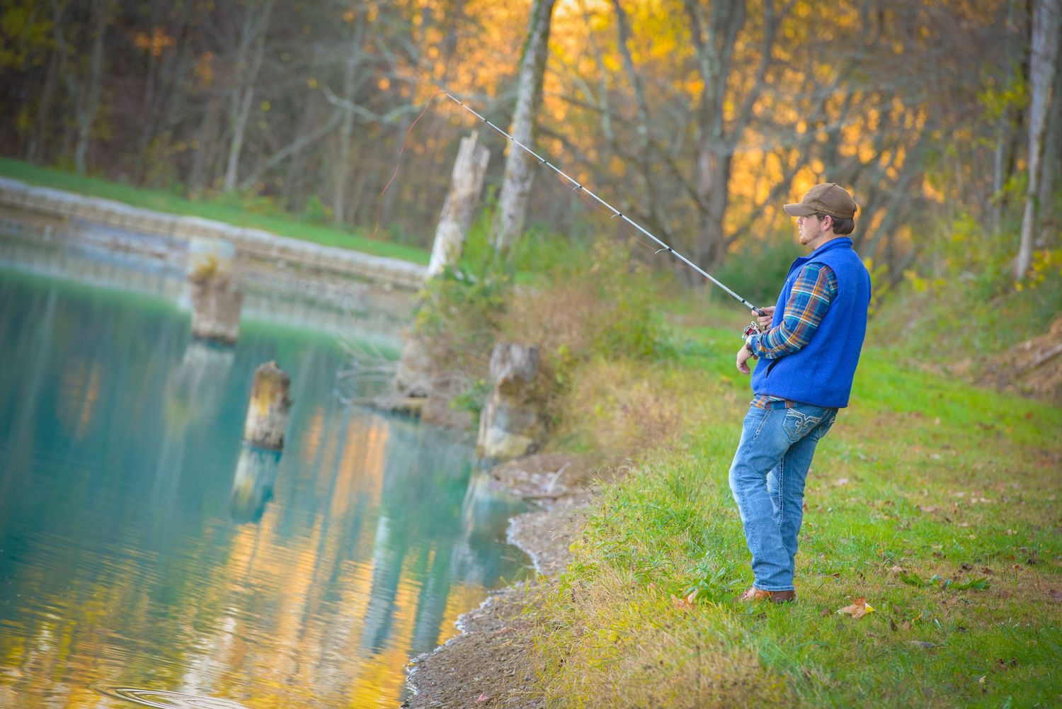 Angler wearing blue jacket fishes at scenic turquoise lake with autumn foliage reflected in calm waters.
