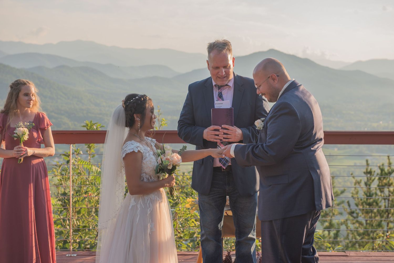 A wedding ceremony takes place on a mountaintop terrace with scenic mountain ranges in the background on a sunny day.
