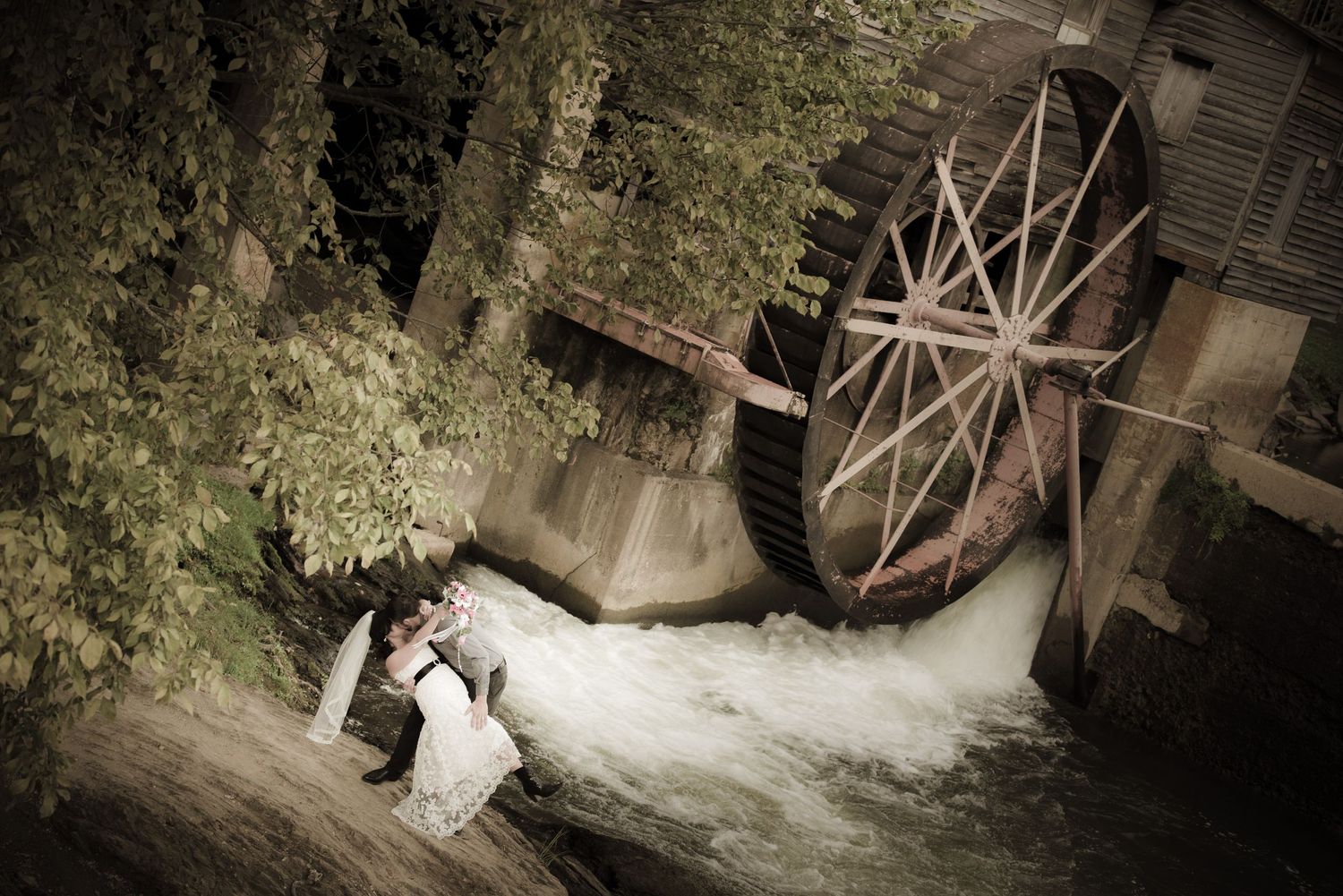 A romantic couple stands near an old water mill wheel with rushing water and lush greenery at Old Mill Pigeon Forge Wedding.
