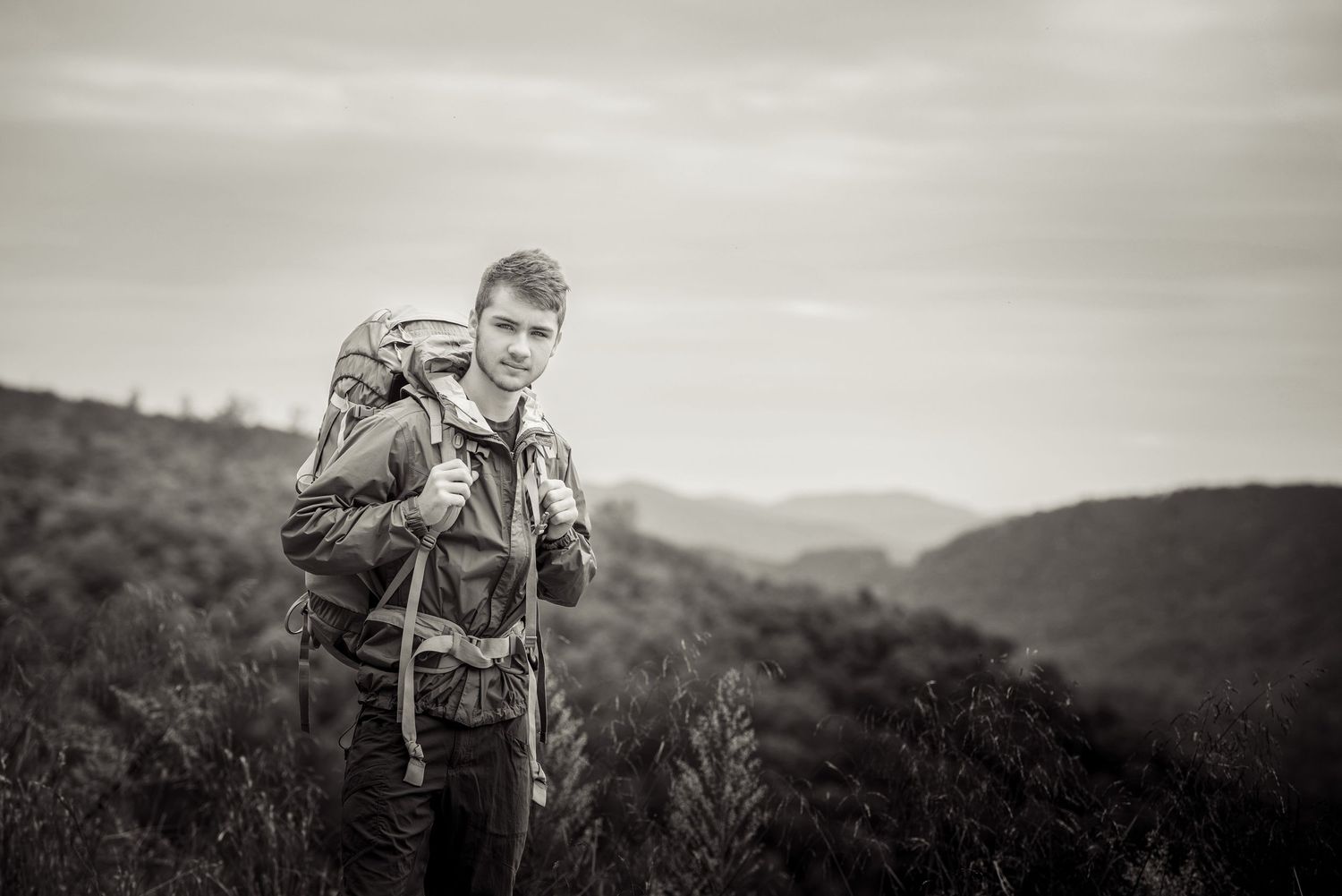 A backpacker with a hiking pack stands on a hillside in black and white against a mountainous landscape.