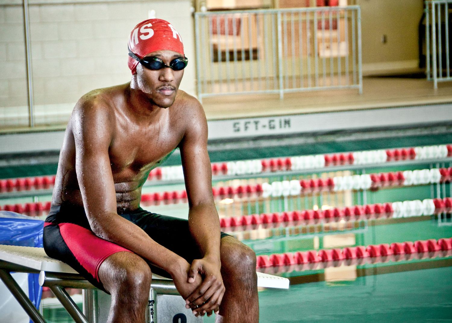 Professional swimmer in red cap and trunks sits at poolside with lane markers visible in background.
