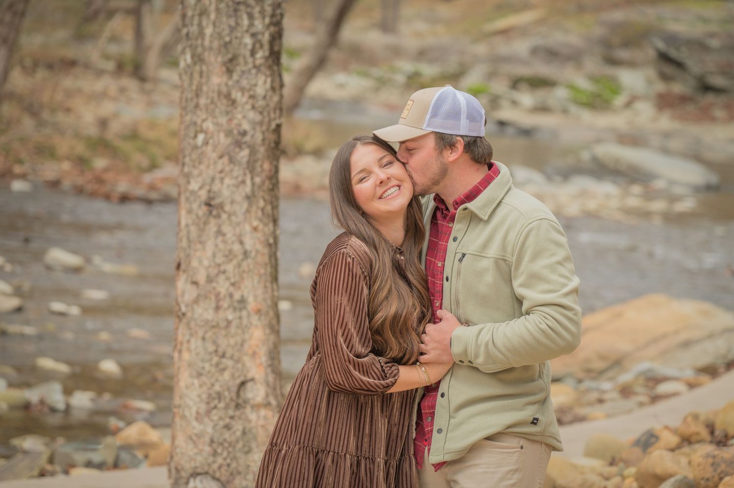 A couple in casual fall attire share a romantic moment near a tree by a rocky stream by Pigeon Forge Photographer tn.