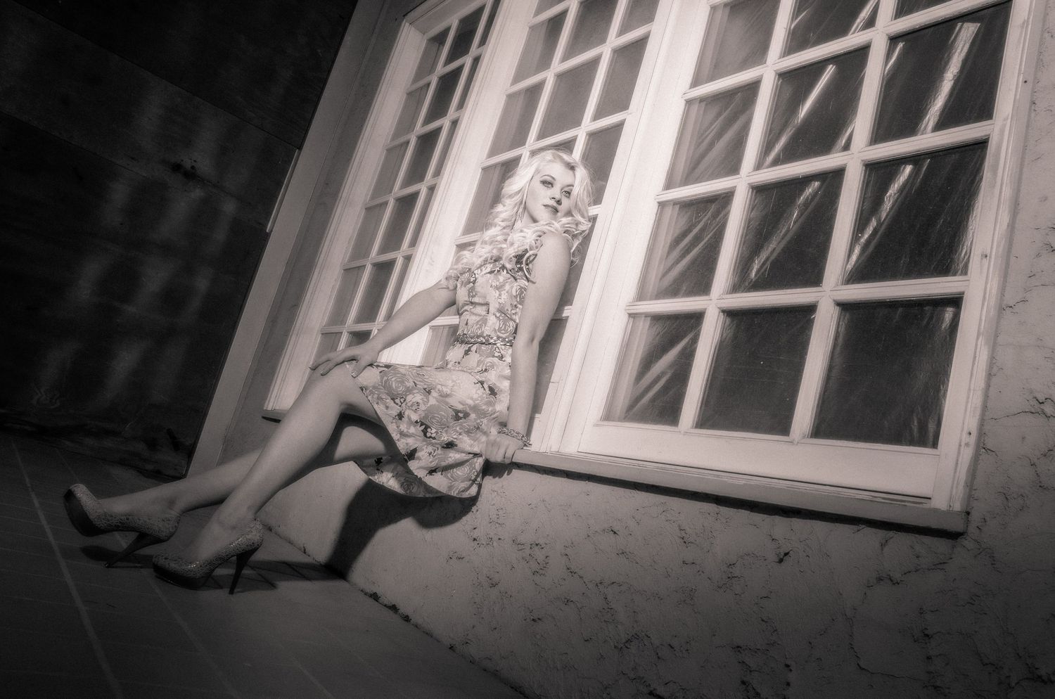 A dark black and white artistic photograph showing a person sitting below a large window in an eerie setting.