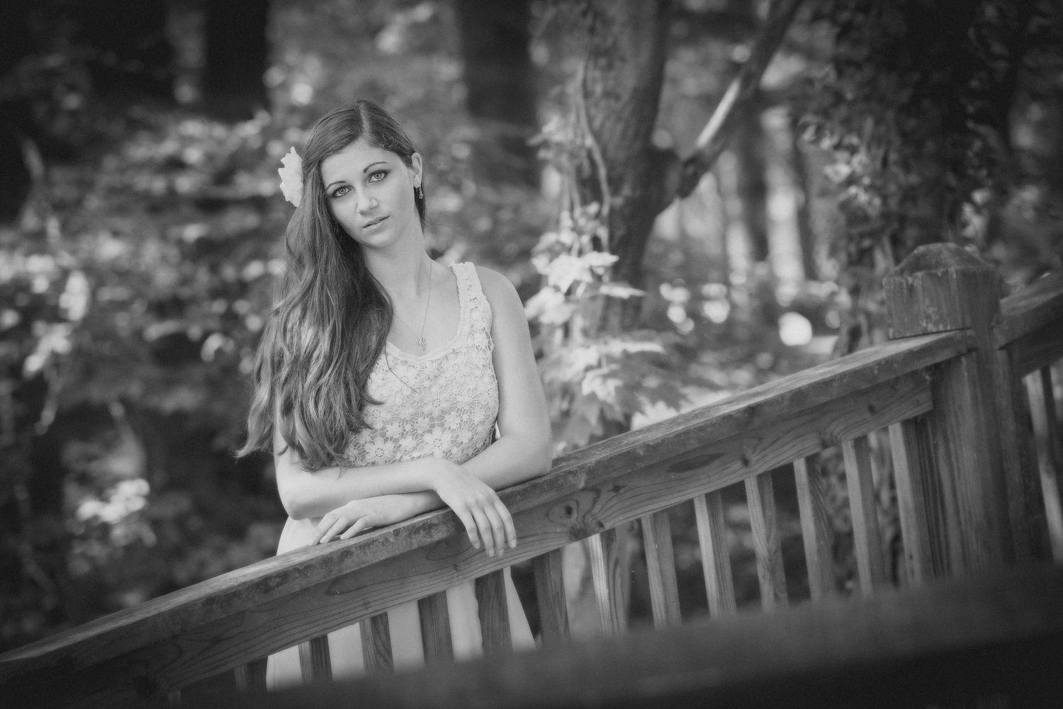 Black and white portrait of someone standing at a wooden railing in a peaceful outdoor forest setting.