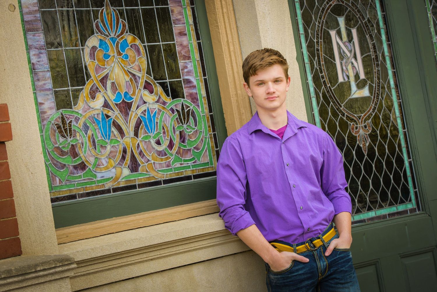 A person in a bright purple shirt leans against a wall with decorative stained glass window and green door.