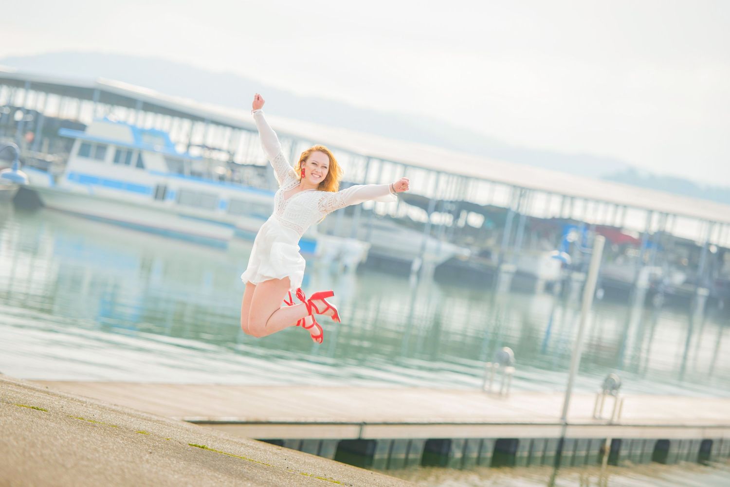 High School Senior Portrait in Dandridge TN of a fun girl jumping in white dress and red shoes against boats dock background in Tennessee.