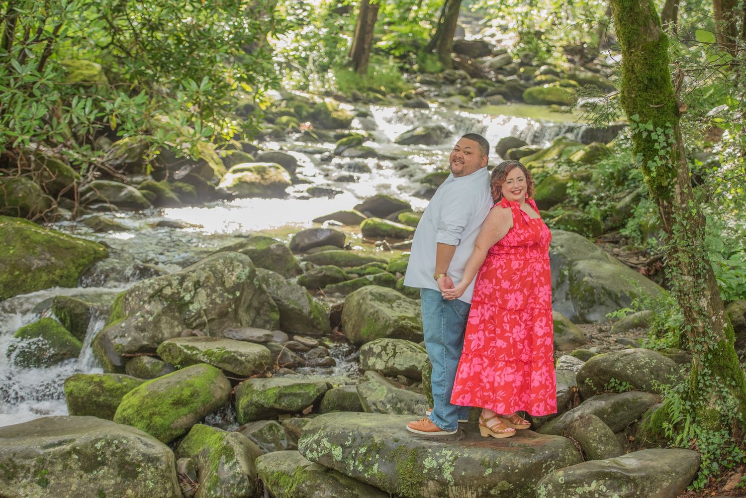 Couple standing together on mossy rocks beside a flowing mountain stream in a lush green forest setting by Sevierville TN Photographers.