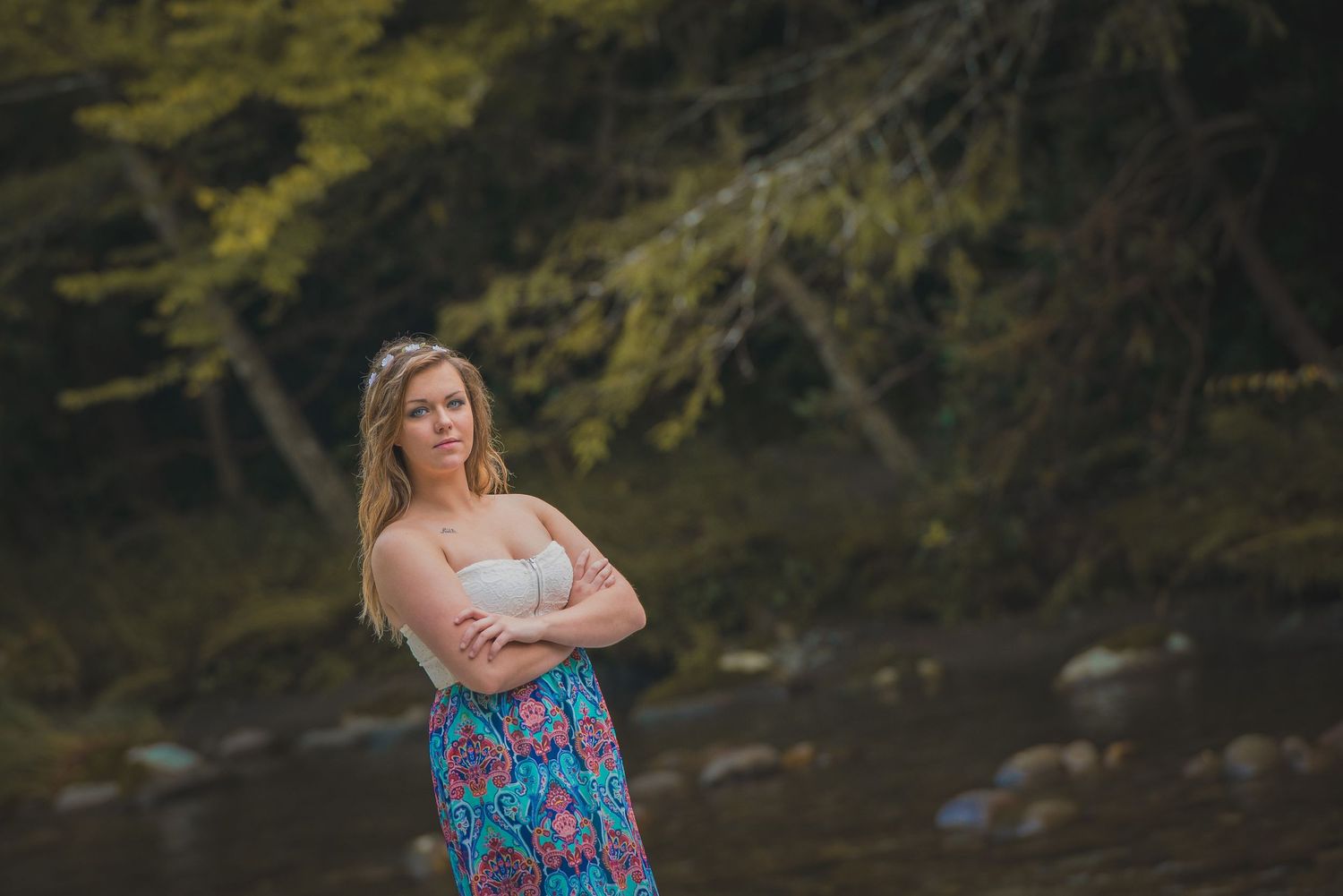 Someone in a floral dress poses with crossed arms by a stream in a wooded setting at dusk.