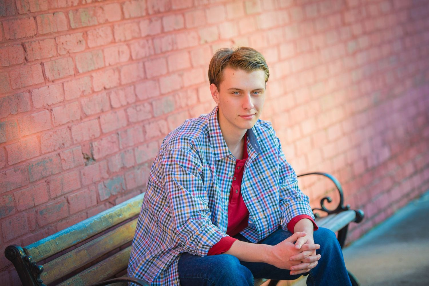 A contemplative portrait of someone in plaid sitting on a bench against a red brick wall.