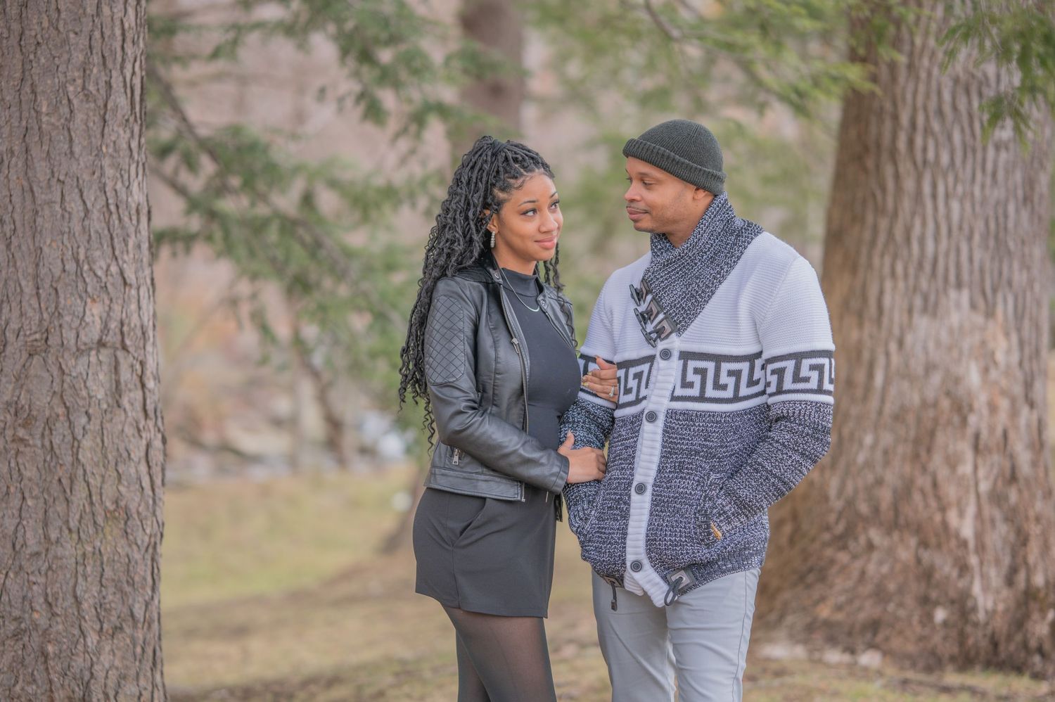 A black couple in warm winter clothing stands close together in a wooded outdoor setting during autumn.