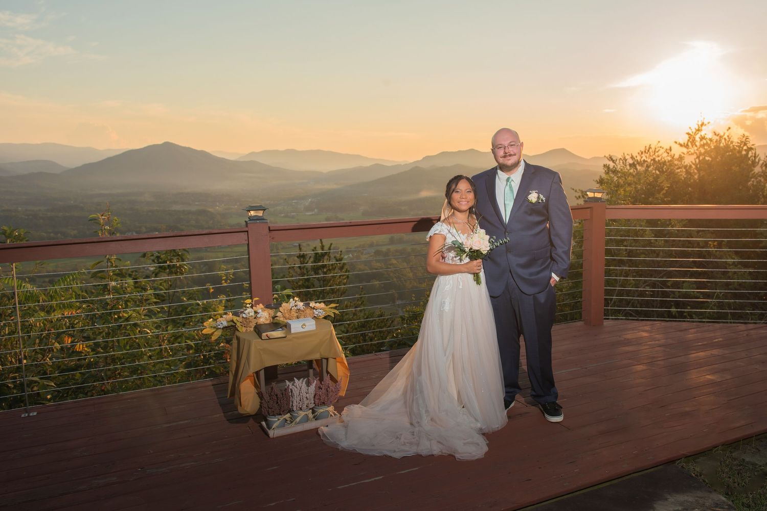 Wedding couple poses on mountain deck overlook during sunset with scenic Blue Ridge Mountains in background.