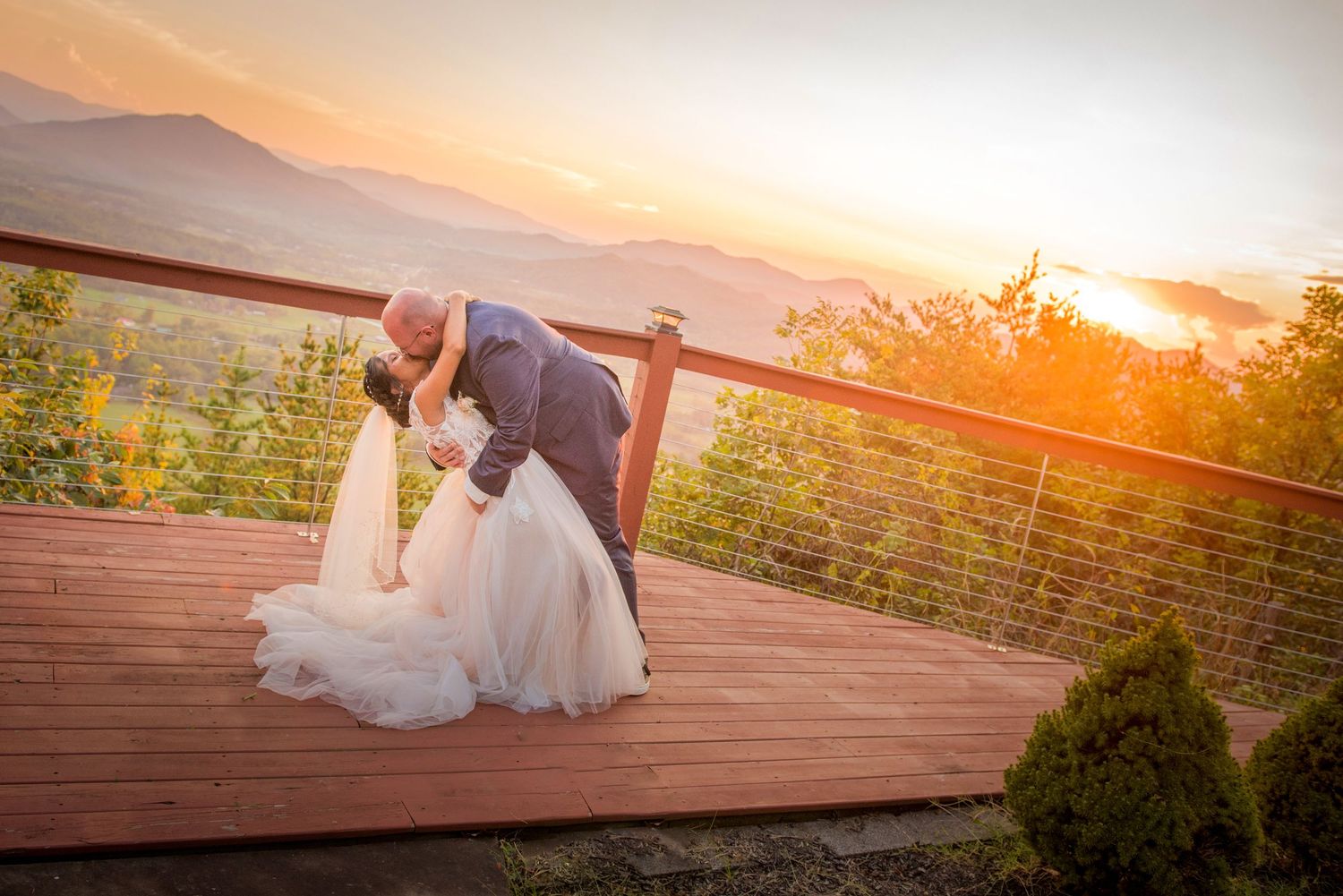 A couple shares a romantic dip kiss on a wooden deck overlooking mountains during a golden sunset by Gatlinburg Wedding Photographers.