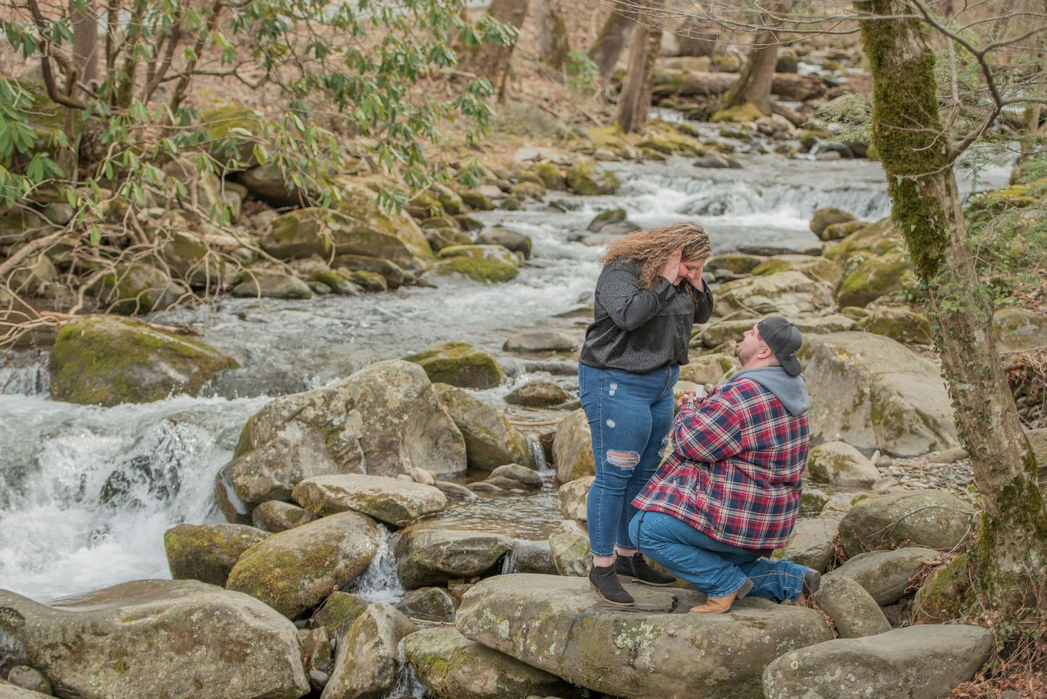 Two people sit on rocks while admiring a cascading mountain stream surrounded by trees and boulders for a surprise proposal in Sevierville.