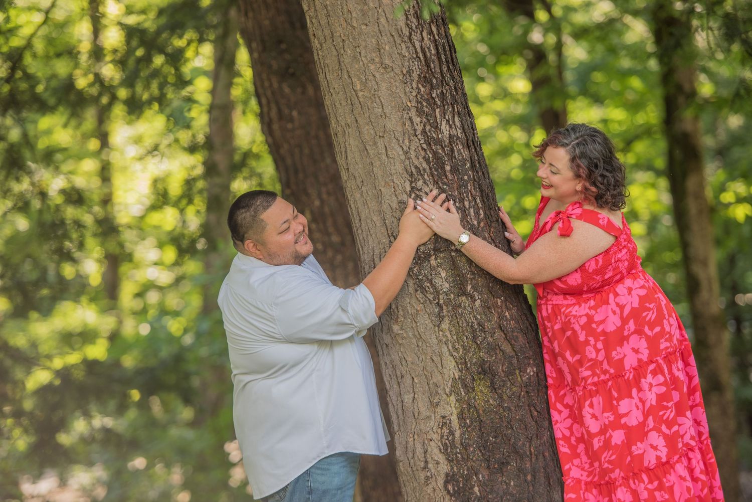 A couple in casual attire playfully poses on opposite sides of a large tree trunk in a sunny wooded setting by Sevierville TN Photographers.