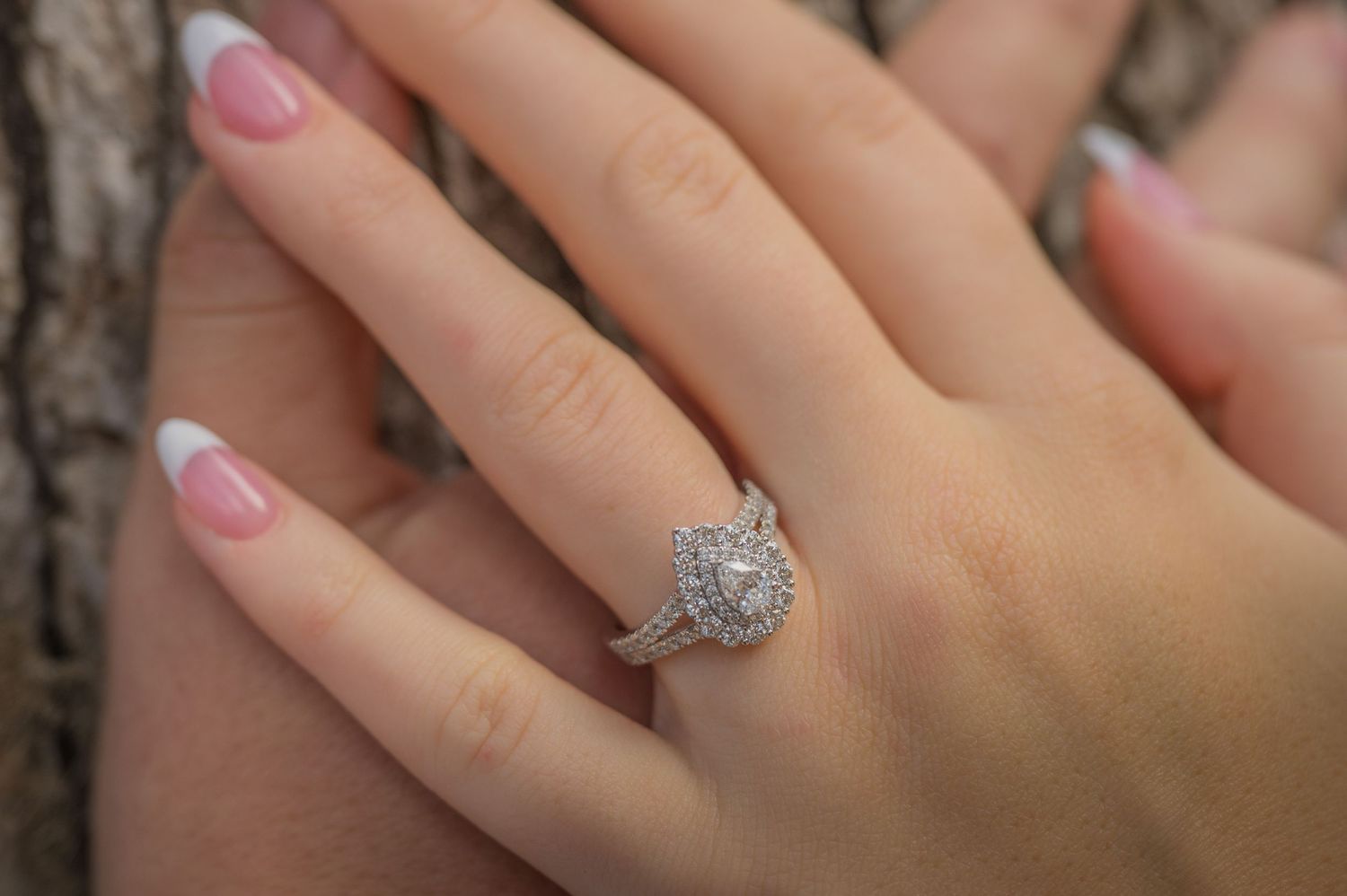 A diamond engagement ring with a round halo design sparkles on a person's hand with pink nail polish.