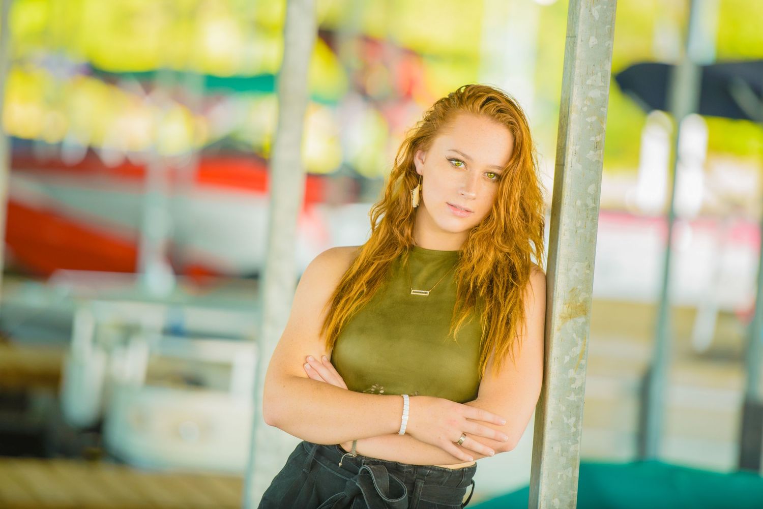 A young woman in an olive green top leans against a pole with blurred colorful background.