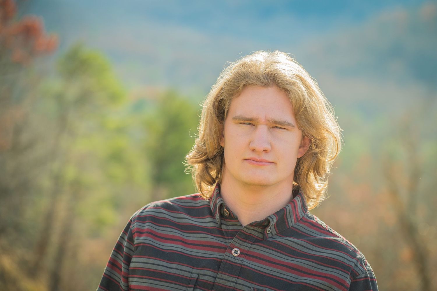 Person with blonde wavy hair wearing a striped shirt poses outdoors against a blurred natural background.