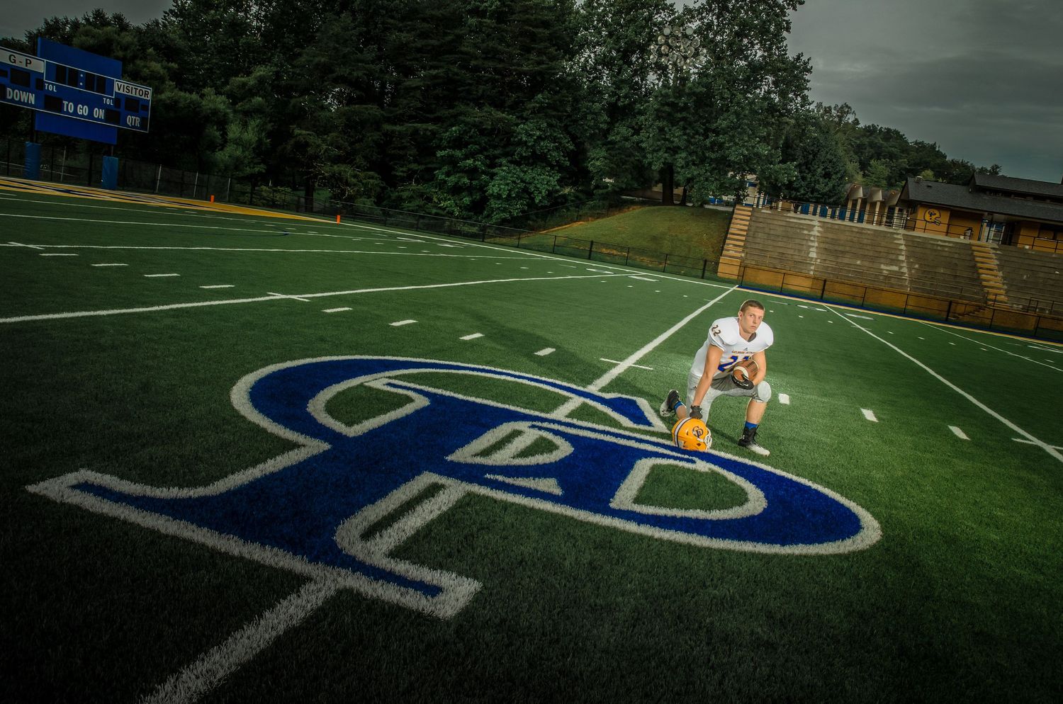 Football player stands on a turf field with a large blue athletic logo painted on the artificial grass surface.