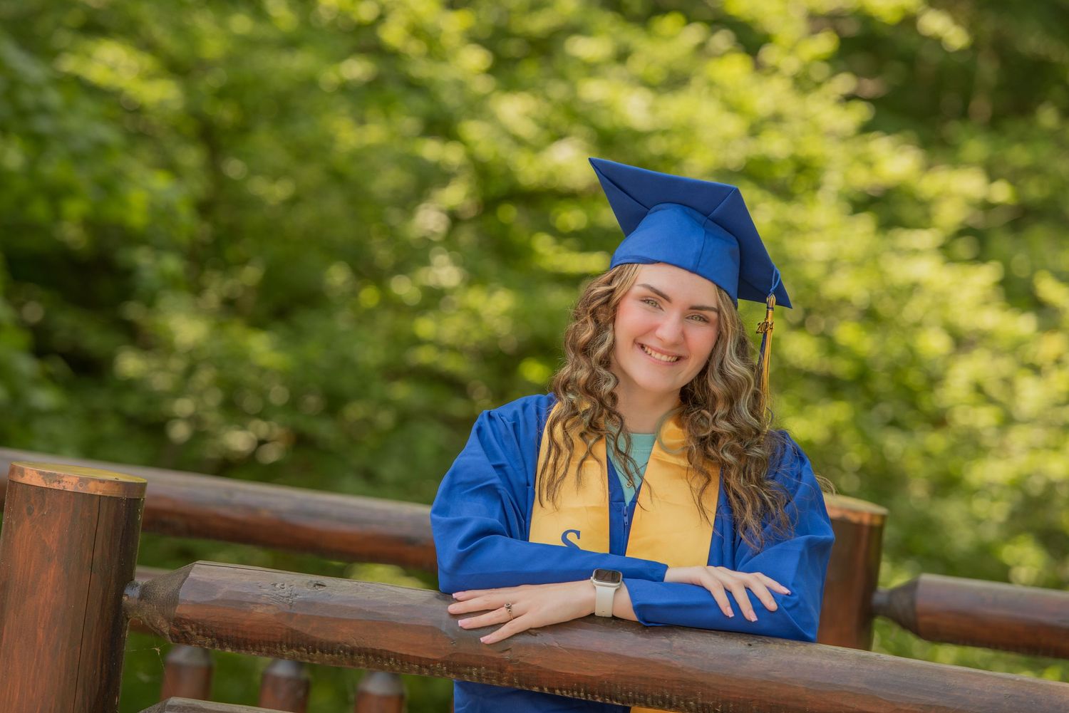 Graduate in bright blue cap and gown with gold stole leans on wooden railing outdoors in natural setting.