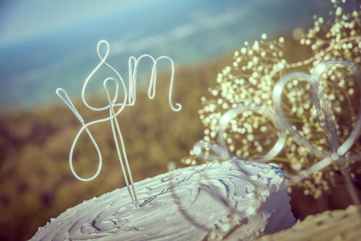 A rustic wedding cake with wire monogram topper and baby's breath flowers set against a blurred outdoor background in Newport TN.